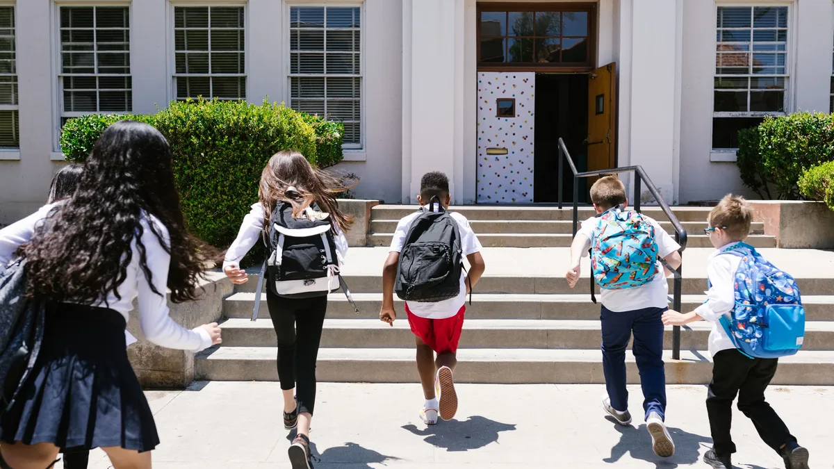 Five children are seen running up the steps of a school building, each carrying a backpack.