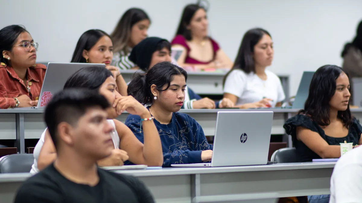 A group of students are seated in a classroom, with some individuals facing forward and others turned to the side, all of whom appear to be of Hispanic or Latin American descent.